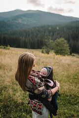 Carrying a baby. Mother with son in sling walk on orange grass in field. Mom, and child in autumn nature. Family with kid hiking. Young tourists on top of a mountain enjoying valley view sunset.