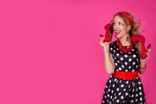  A Blonde Girl In A Black Dress With White Polka Dots With Red Accessories Stands On A Pink Background Holding High-heeled Shoes In Her Hands. Pin-up