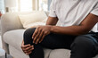Knee pain, hand of a man and injury while sitting on home living room sofa for rest and relaxation. Ache, painful leg joint and african american man touching his knees for a strain muscle