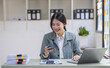 © David - Smiling Asian business woman at work Using  phone and working on laptop sitting at her working place in office, copy space