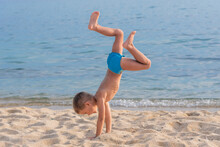 A Child On The Beach Does Sports And Performs Gymnastic Exercises Against The Background Of The Sea.  A Child Does A Handstand On The Beach