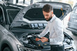 © standret - Looking under the hood and holding tablet. Young man in white clothes is in the car dealership