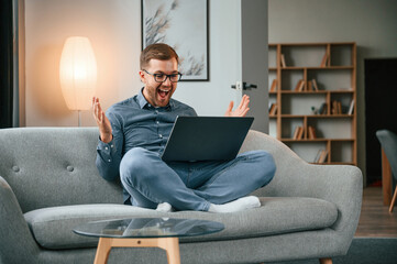 Wall Mural - Celebrating success. Male freelance worker is using laptop at home. Cozy apartment