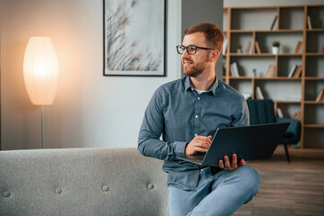 Wall Mural - Sitting and looking at the side. Male freelance worker is using laptop at home. Cozy apartment