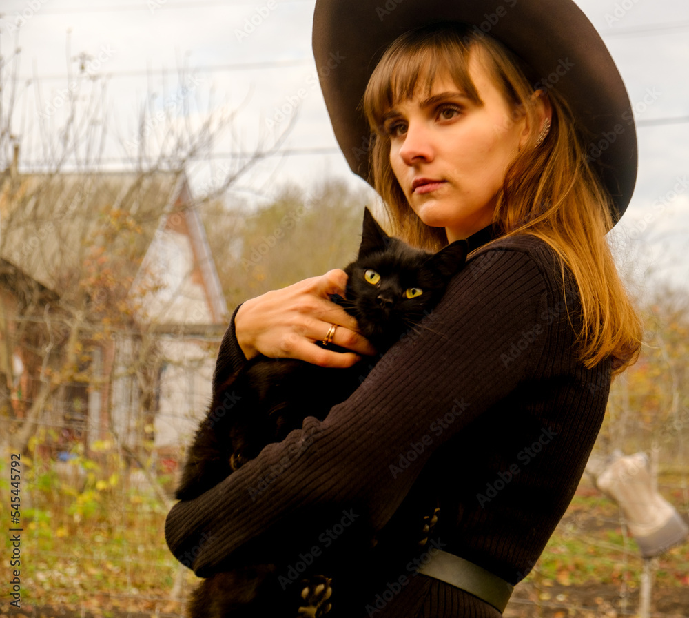 Beautiful young girl in a black dress and in a hat with a black cat ...