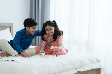 Wall Mural - Happy Indian family, brother and sister with traditional clothes playing fun game together, two children boy and girl sitting talking on bed at home, sibling relationship concept