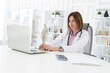 © Vitaliy - Portrait photo of young and beautiful smiling female doctor in white medical gown sitting and working on laptop in the office of the modern clinic. Online consultation and distant cure concept