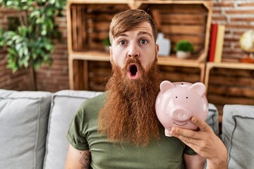 Poster - Redhead man holding piggy bank scared and amazed with open mouth for surprise, disbelief face