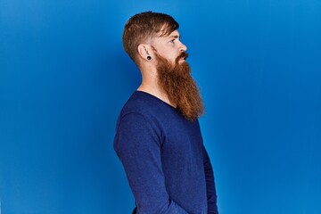 Poster - Redhead man with long beard wearing casual blue sweater over blue background looking to side, relax profile pose with natural face and confident smile.