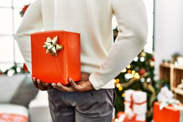 Poster - African american man holding gift on his back standing by christmas tree at home.