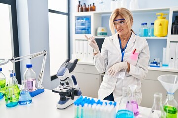 Poster - Middle age blonde woman working at scientist laboratory pointing aside worried and nervous with forefinger, concerned and surprised expression