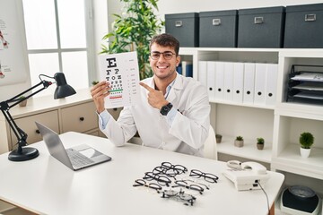 Poster - Young hispanic man working at ophthalmology clinic smiling happy pointing with hand and finger