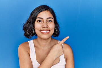 Sticker - Young hispanic woman standing over blue background cheerful with a smile of face pointing with hand and finger up to the side with happy and natural expression on face