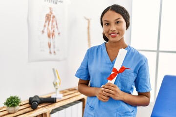 Wall Mural - Young latin woman wearing physiotherapist uniform holding diploma at clinic