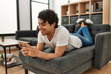 Poster - Young hispanic man using smartphone lying on the sofa at home.