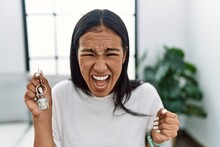 Young Hispanic Woman Holding Keys Of New Home Angry And Mad Screaming Frustrated And Furious, Shouting With Anger. Rage And Aggressive Concept.