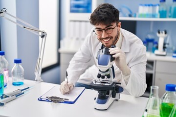 Wall Mural - Young hispanic man scientist writing on document using microscope at laboratory