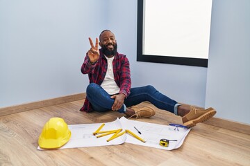 Sticker - African american man sitting on the floor at new home looking at blueprints smiling looking to the camera showing fingers doing victory sign. number two.