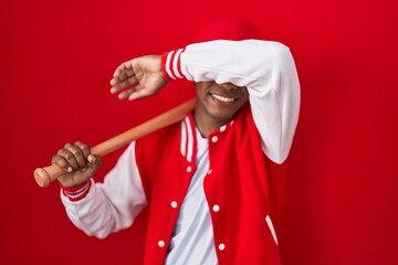 Wall Mural - Young hispanic man playing baseball holding bat smiling cheerful playing peek a boo with hands showing face. surprised and exited