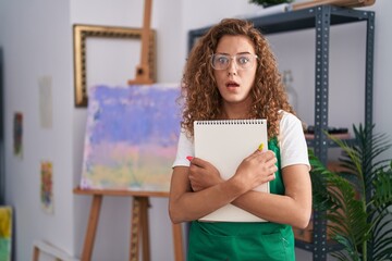 Sticker - Young caucasian woman holding art notebook afraid and shocked with surprise and amazed expression, fear and excited face.