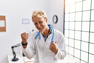 Poster - Young blond man wearing doctor uniform and stethoscope at clinic very happy and excited doing winner gesture with arms raised, smiling and screaming for success. celebration concept.