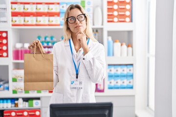 Sticker - Young blonde woman working at pharmacy drugstore holding paper bag serious face thinking about question with hand on chin, thoughtful about confusing idea