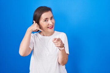 Wall Mural - Middle age hispanic woman standing over blue background smiling doing talking on the telephone gesture and pointing to you. call me.