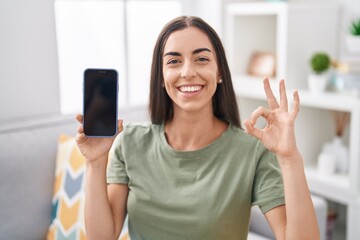 Poster - Young brunette woman holding smartphone showing blank screen doing ok sign with fingers, smiling friendly gesturing excellent symbol