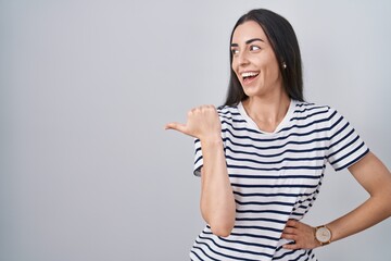 Wall Mural - Young brunette woman wearing striped t shirt smiling with happy face looking and pointing to the side with thumb up.