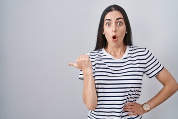 Wall Mural - Young brunette woman wearing striped t shirt surprised pointing with hand finger to the side, open mouth amazed expression.