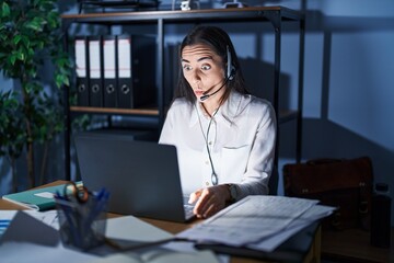 Poster - Young brunette woman wearing call center agent headset working late at night making fish face with lips, crazy and comical gesture. funny expression.