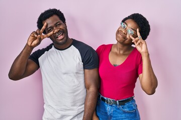 Poster - Young african american couple standing over pink background doing peace symbol with fingers over face, smiling cheerful showing victory