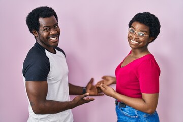 Poster - Young african american couple standing over pink background inviting to enter smiling natural with open hand