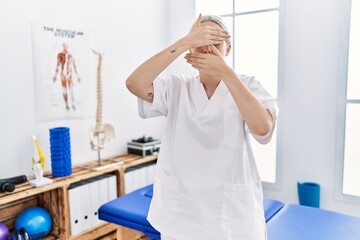 Wall Mural - Young caucasian woman working at pain recovery clinic covering eyes and mouth with hands, surprised and shocked. hiding emotion