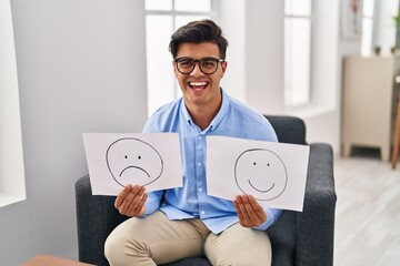 Wall Mural - Hispanic man working on depression holding sad to happy emotion paper smiling and laughing hard out loud because funny crazy joke.