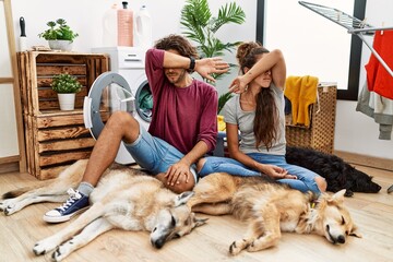 Poster - Young hispanic couple doing laundry with dogs covering eyes with arm, looking serious and sad. sightless, hiding and rejection concept