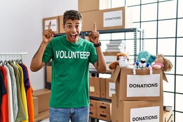 Poster - Young handsome hispanic man wearing volunteer t shirt at donations stand smiling amazed and surprised and pointing up with fingers and raised arms.