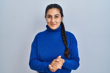 Poster - Young hispanic woman standing over isolated background with hands together and crossed fingers smiling relaxed and cheerful. success and optimistic