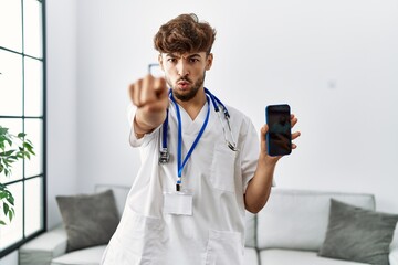 Canvas Print - Young arab man wearing doctor uniform and stethoscope holding smartphone pointing with finger to the camera and to you, confident gesture looking serious