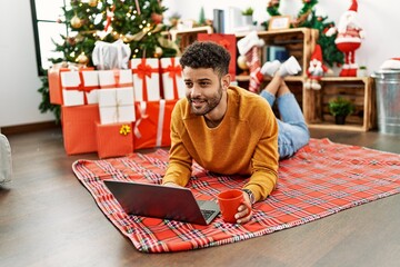 Canvas Print - Young arab man using laptop and drinking coffee lying on the floor by christmas tree at home.