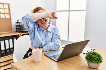 Canvas Print - Young redhead woman working at the office using computer laptop covering eyes with arm smiling cheerful and funny. blind concept.