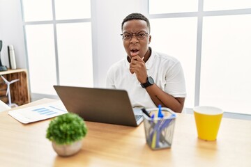 Wall Mural - Young african man working at the office using computer laptop looking fascinated with disbelief, surprise and amazed expression with hands on chin