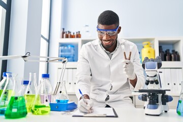 Canvas Print - Young african american man wearing scientist uniform writing on clipboard holding test tubes at laboratory