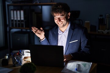 Poster - Hispanic young man working at the office at night smiling cheerful presenting and pointing with palm of hand looking at the camera.