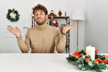 Poster - Young handsome man with beard sitting on the table by christmas decoration clueless and confused expression with arms and hands raised. doubt concept.