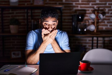 Canvas Print - Hispanic man with beard using laptop at night shocked covering mouth with hands for mistake. secret concept.