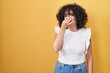 Young middle east woman standing over yellow background smelling something stinky and disgusting, intolerable smell, holding breath with fingers on nose. bad smell