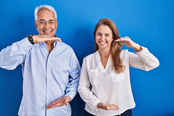 Wall Mural - Middle age hispanic couple standing over blue background gesturing with hands showing big and large size sign, measure symbol. smiling looking at the camera. measuring concept.