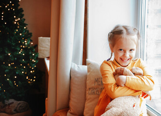 Wall Mural - A cute preschool girl sitting on the windowsill with her plush toy waiting for Christmas near the christmas tree. Winter holidays concept