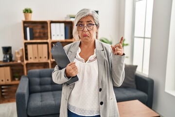 Poster - Middle age woman with grey hair at consultation office pointing up looking sad and upset, indicating direction with fingers, unhappy and depressed.